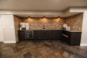 Indoor wet bar featuring dark wood finish cabinetry, dark stone countertops, beverage cooler, a textured ceiling, and brick wall