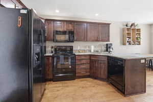 Kitchen featuring black appliances, dark wood finish cabinets, a peninsula, light stone counters, and recessed lighting
