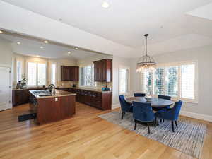 Kitchen with an island with sink, light wood finished floors, light stone countertops, dark wood finish cabinetry, and vaulted ceiling
