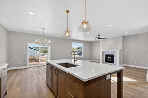 Kitchen featuring ceiling fan, a glass covered fireplace, light wood-style flooring, light stone countertops, and an island with sink