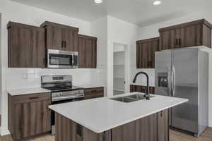 Kitchen featuring stainless steel appliances, dark wood finish cabinets, backsplash, an island with sink, and light wood finished floors