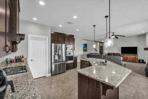Kitchen featuring dark wood finish cabinets, light stone countertops, a ceiling fan, stainless steel appliances, and open floor plan