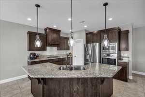 Kitchen featuring dark wood finish cabinetry, light stone counters, stainless steel appliances, a kitchen breakfast bar, and a kitchen island with sink