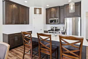 Kitchen with dark wood finish cabinets, stainless steel appliances, decorative light fixtures, dark wood-style floors, and an island with sink
