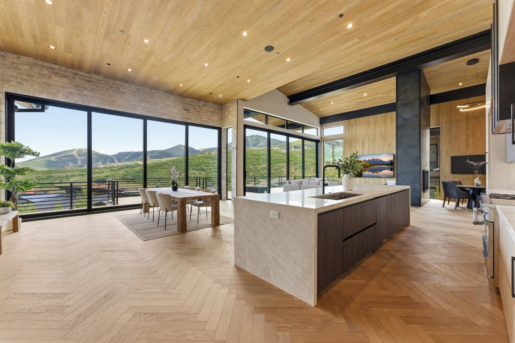 Expansive interior featuring floor-to-ceiling windows, wood-finish herringbone flooring, a large kitchen island with integrated sink, exposed structural beams, and a wood-paneled ceiling