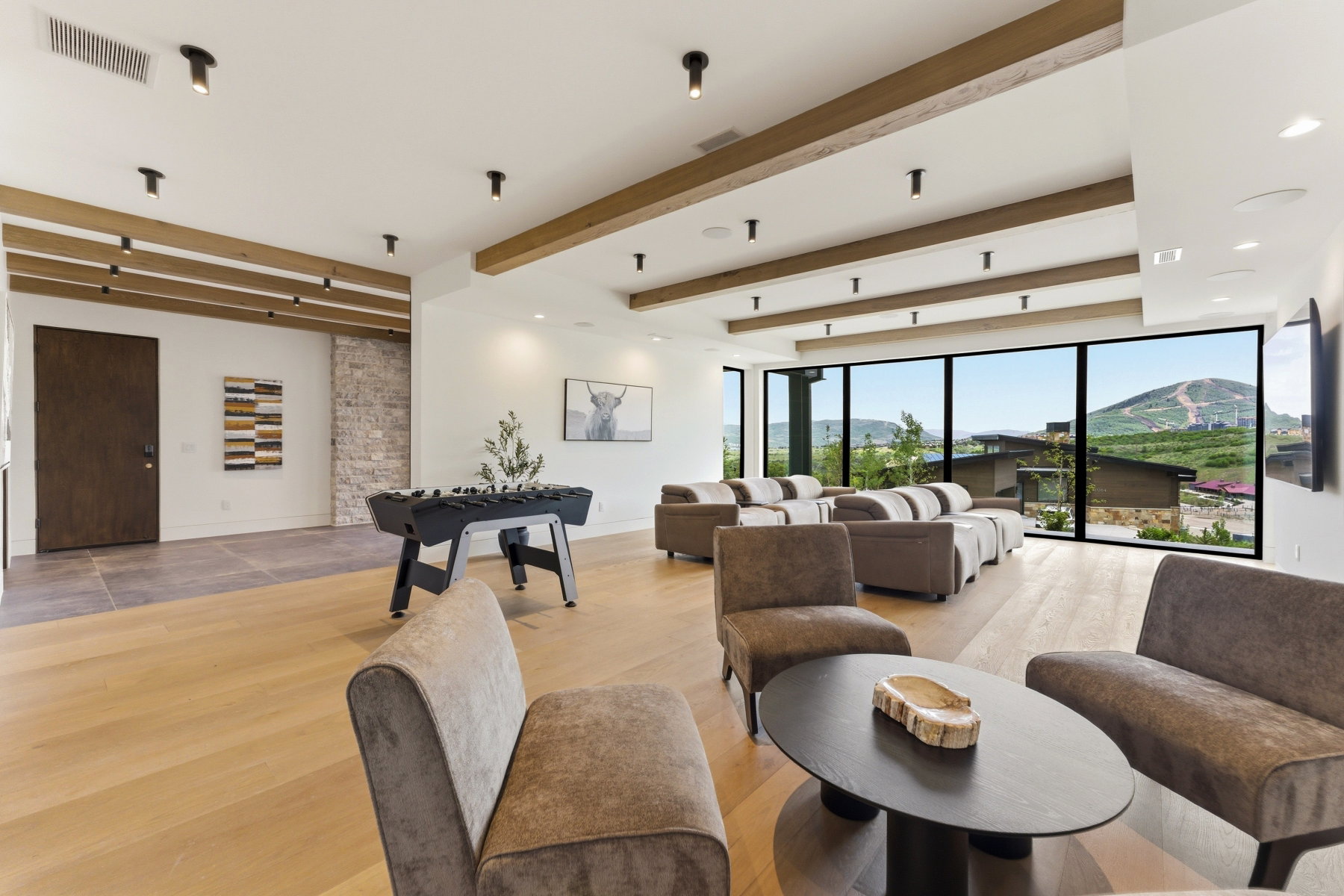 Spacious room featuring light wood-finish flooring, exposed wood ceiling beams, and extensive floor-to-ceiling windows