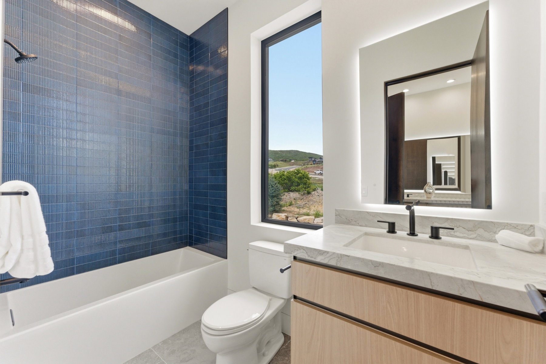 Bathroom featuring a built-in tub with blue subway tile surround, a single sink vanity with a light wood-finish cabinet, a light-toned countertop, and a back-lit mirror