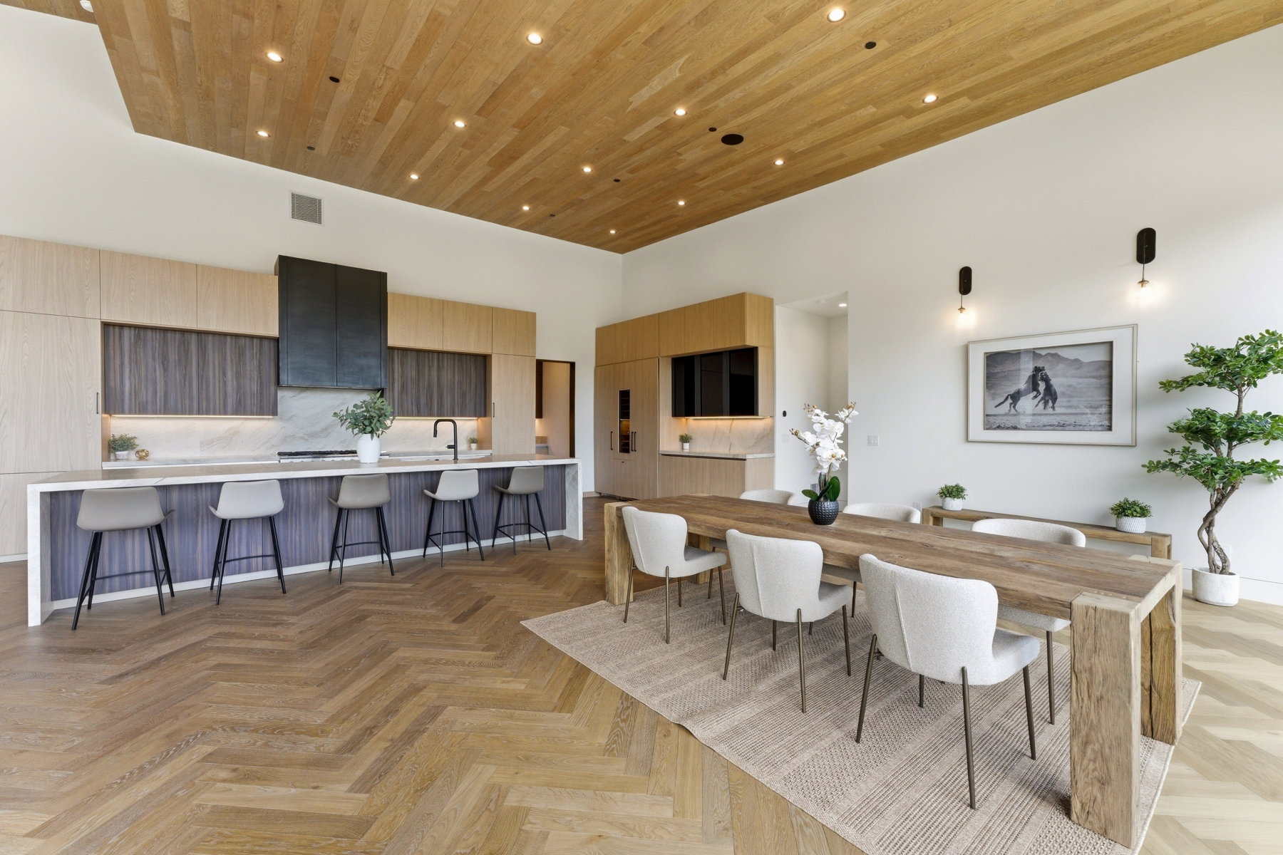 Expansive open-concept space featuring herringbone wood-finish flooring, a light wood-paneled ceiling with recessed lighting, and a contemporary kitchen with an elongated island, light-toned cabinetry, and a dark range hood