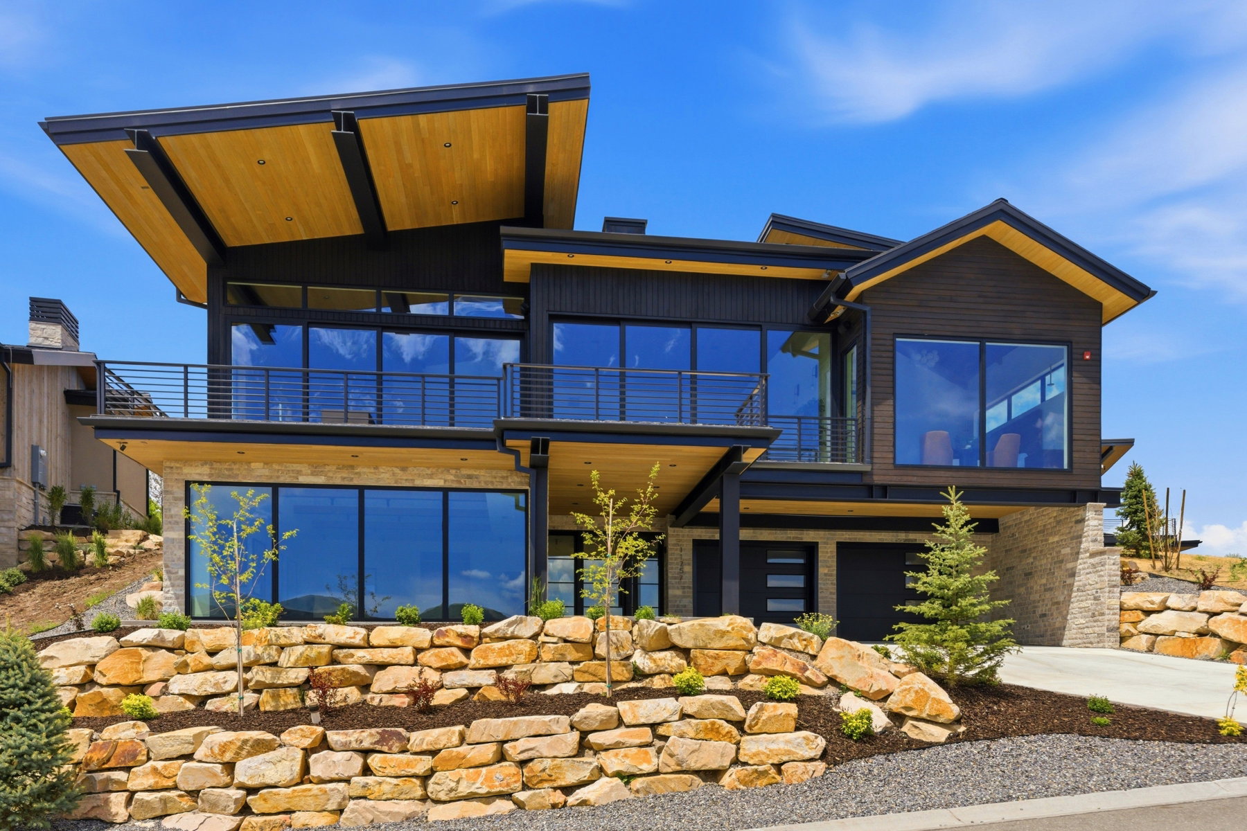 Modern architectural exterior featuring multiple expansive windows, a spacious balcony with horizontal railings, a prominent wood-finish soffit, stone retaining walls, and a multi-level design