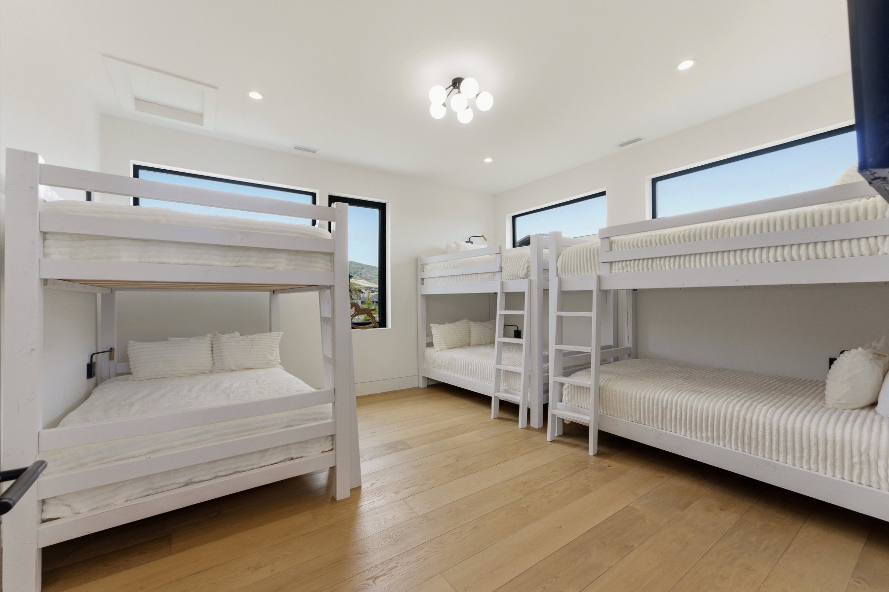 Bright room with light wood-finish flooring, featuring multiple white bunk beds, recessed lighting, and a contemporary ceiling fixture