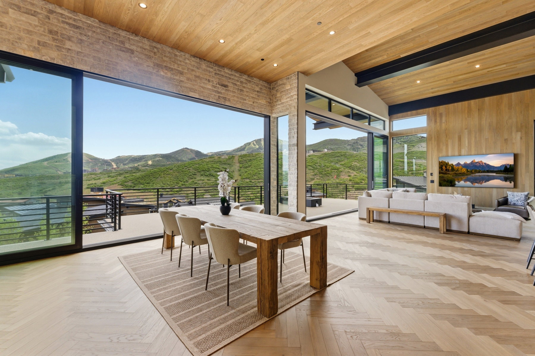Expansive great room featuring a pitched wood-clad ceiling with exposed beams, herringbone wood-finish flooring, a full-height stone accent wall, and floor-to-ceiling glass paneling opening to an outdoor deck with mountain scenery