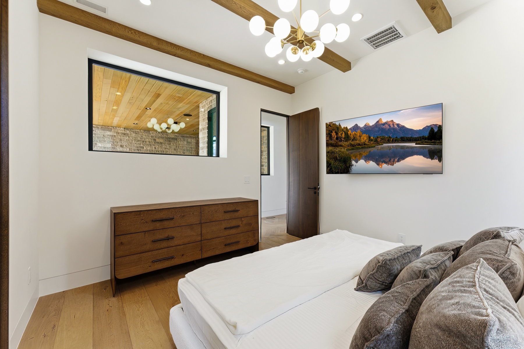 Bedroom featuring exposed ceiling beams, a contemporary chandelier, light wood-finish flooring, and a large horizontal window providing a unique interior perspective