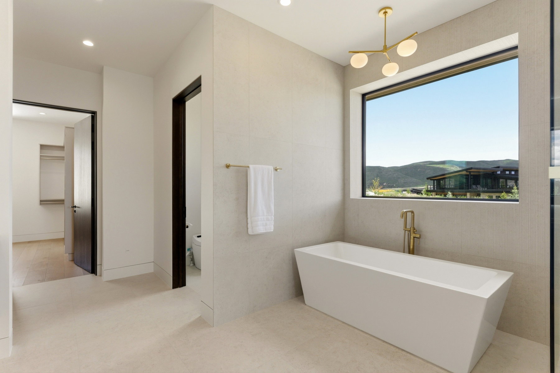 Modern bathroom featuring a freestanding soaking tub with brass fixtures, a large picture window, and a contemporary brass chandelier