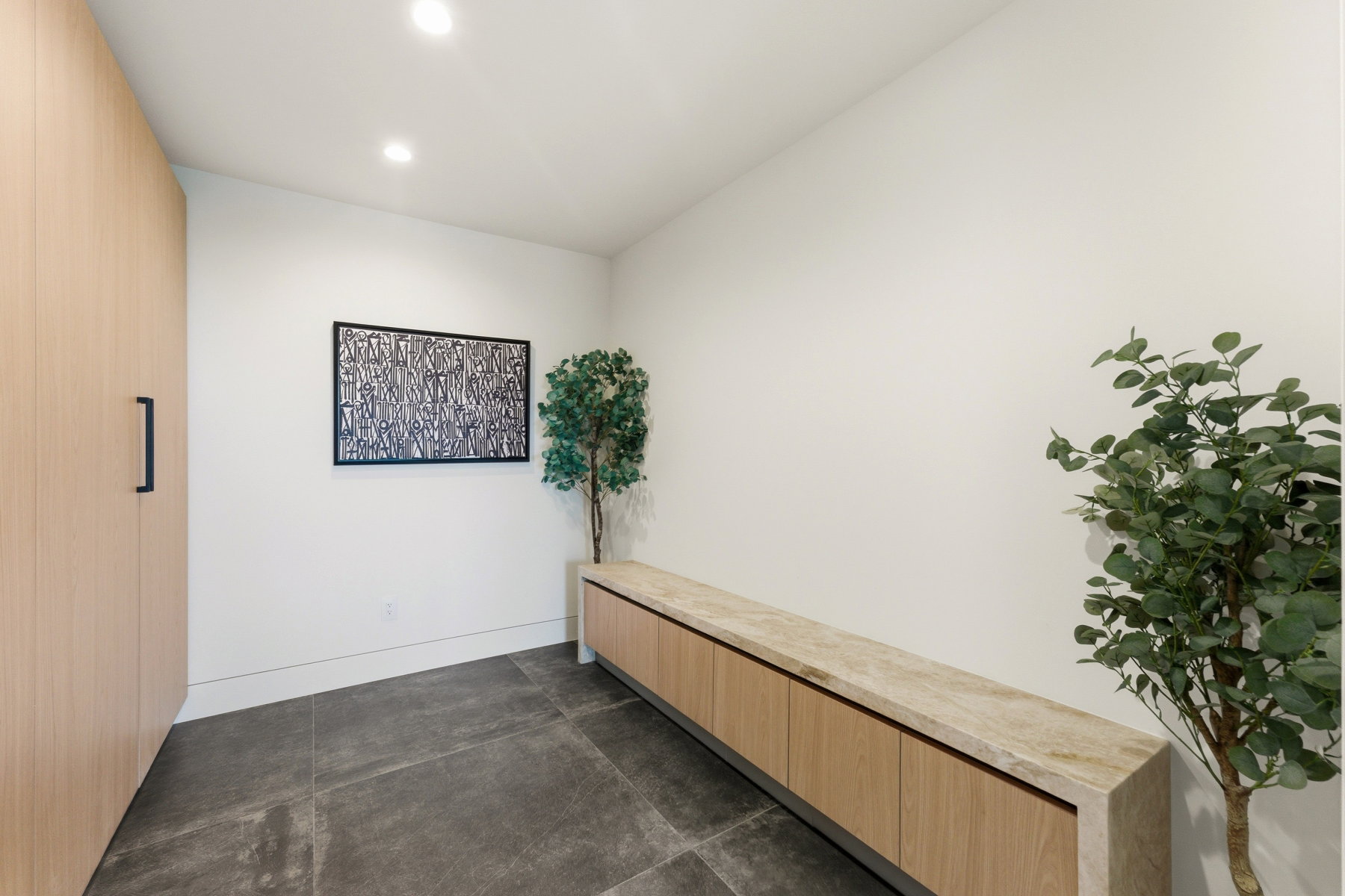 Recessed lighting illuminating a space featuring a floor-to-ceiling wood-finish storage unit with black hardware, and dark large-format floor tiles