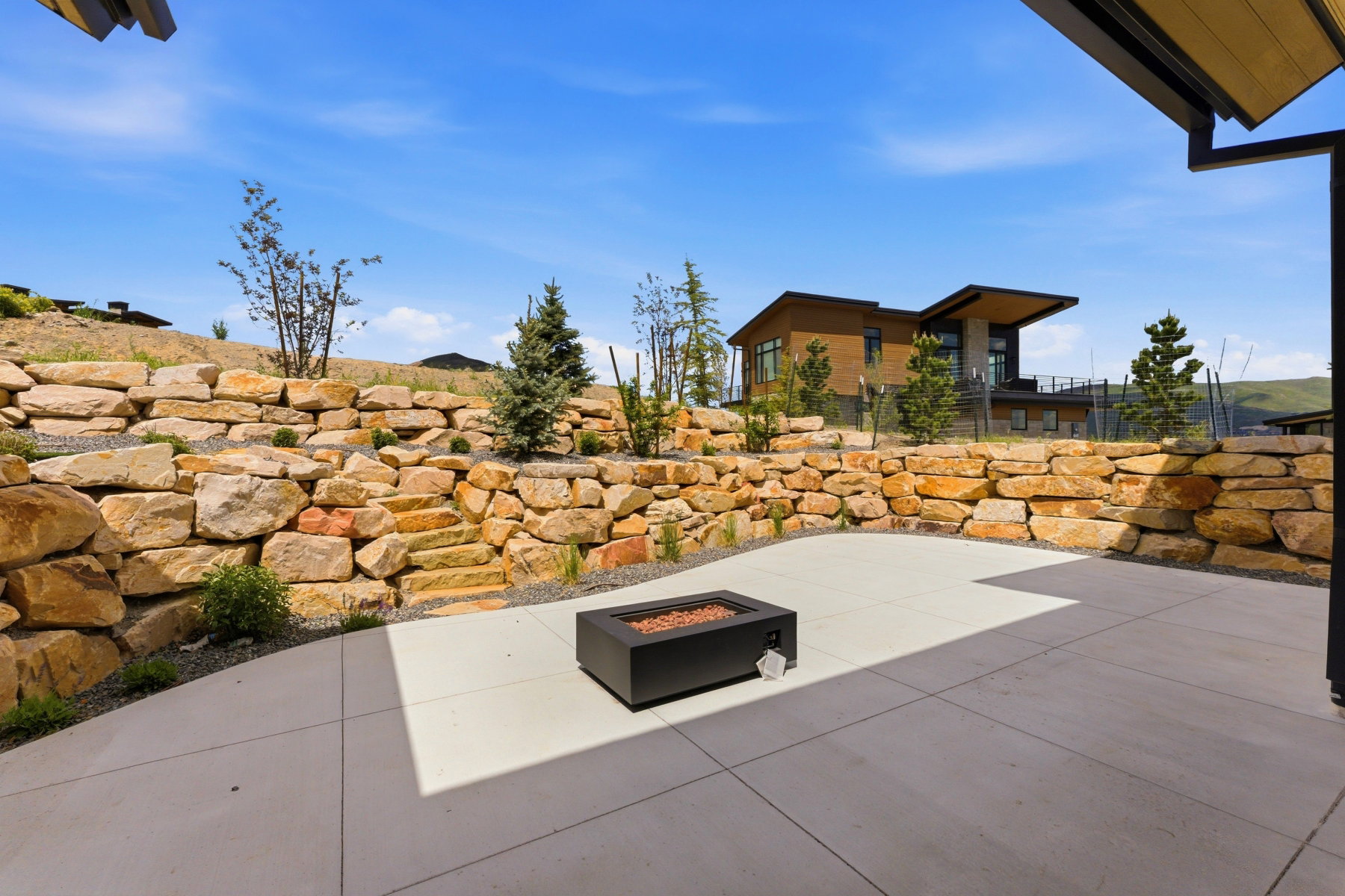 Spacious concrete patio with a modern fire pit, surrounded by a natural stone retaining wall