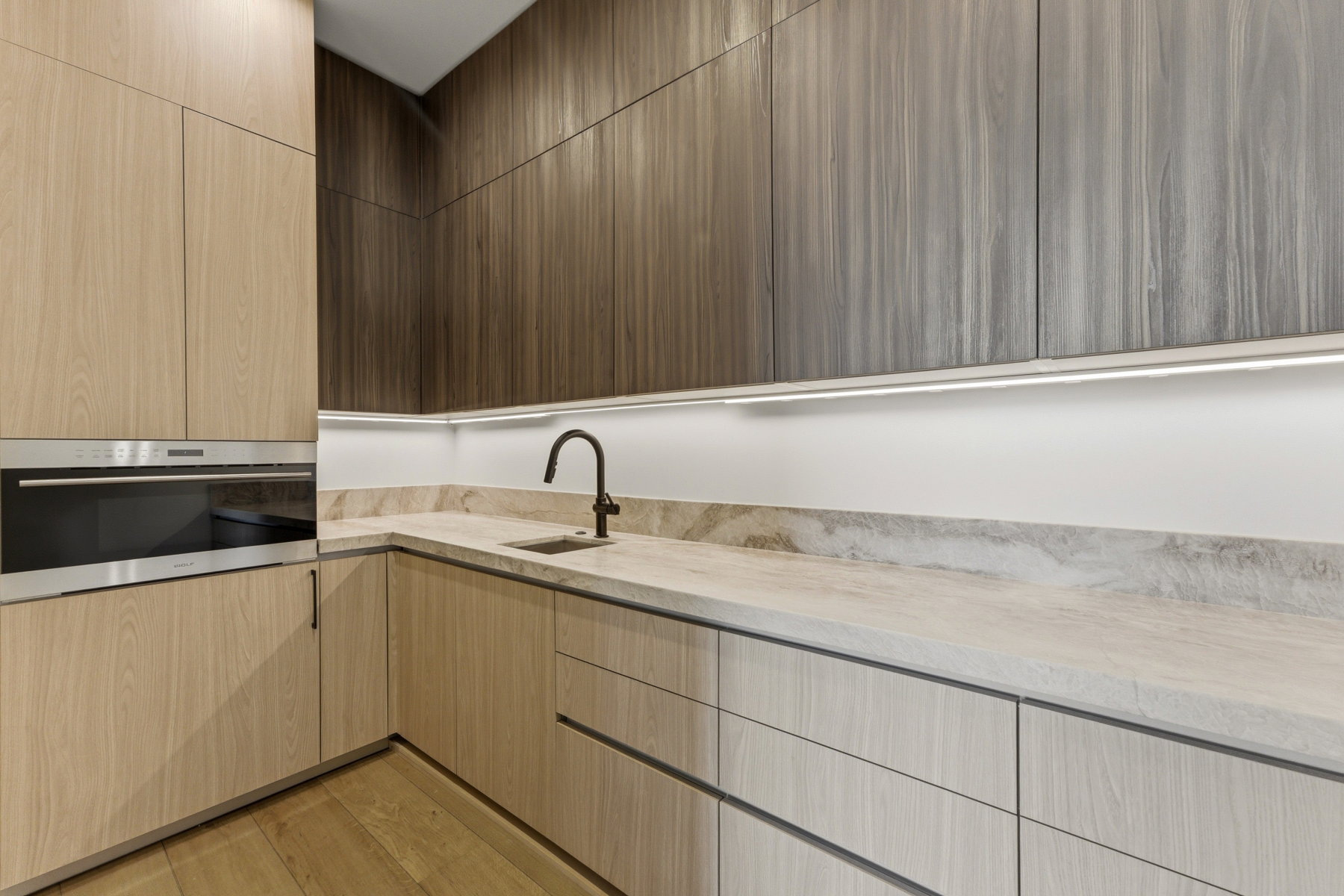 Contemporary kitchen featuring two-tone wood-finish cabinetry, integrated stainless steel oven, undermount sink with a matte black faucet, and stone countertops