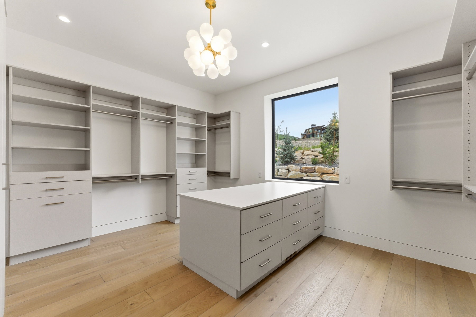 Spacious walk-in closet featuring light wood-finish flooring, built-in shelving, ample hanging rods, and a central island with integrated drawers and a light-colored countertop