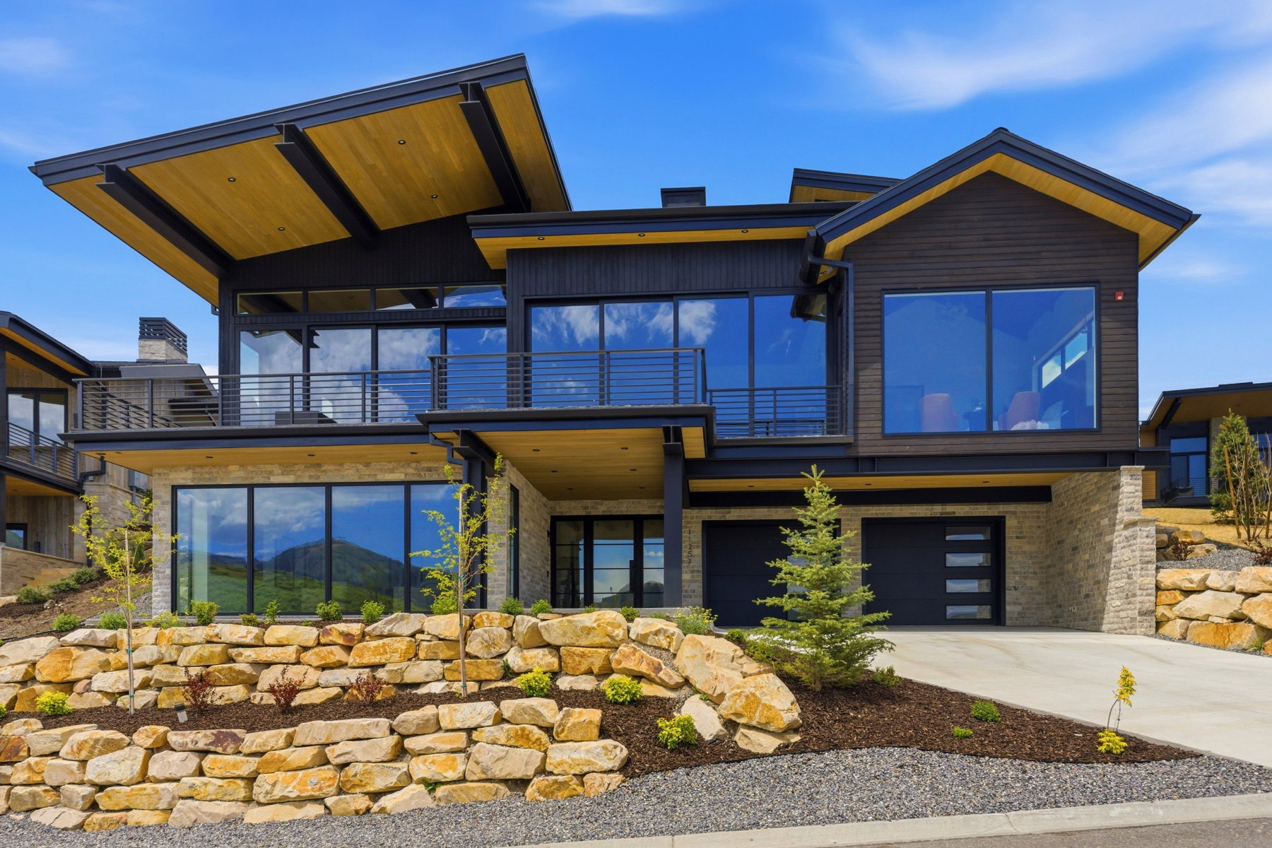 Modern multi-level residence featuring expansive window walls, a striking wood-clad overhang, and a stone retaining wall