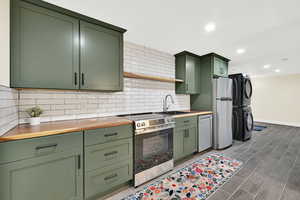 Kitchen with green cabinetry, stainless steel appliances, wood counters, stacked washing machine and dryer, and recessed lighting