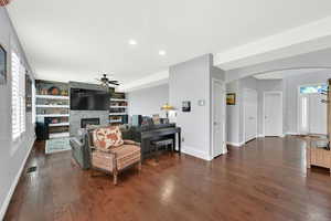 Living area featuring arched walkways, dark wood-type flooring, a stone fireplace, ceiling fan, and recessed lighting