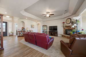 Living room featuring a raised ceiling, arched walkways, a fireplace, a ceiling fan, and light wood-style flooring