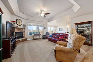 Living room with a tray ceiling, ceiling fan, a stone fireplace, hanging lights, and light colored carpet