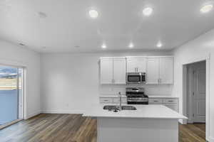 Kitchen with stainless steel appliances, dark wood-style floors, white cabinets, a center island with sink, and light stone countertops