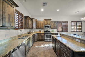 Kitchen featuring dark wood finish cabinets, light stone countertops, stainless steel appliances, suspended lighting, and a center island with sink