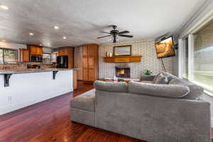 Living room with dark wood-style flooring, a brick fireplace, a ceiling fan, a textured ceiling, and brick wall