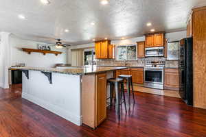 Kitchen with a kitchen breakfast bar, stainless steel appliances, wood finish cabinets, dark wood-style flooring, and recessed lighting
