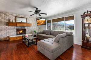 Living room with brick wall, dark wood-style floors, a brick fireplace, a textured ceiling, and ceiling fan
