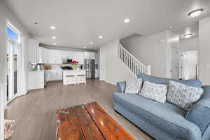 Living room with light wood-style flooring and recessed lighting, looking into the kitchen with stainless steel appliances, white cabinetry, light stone countertops, light wood-style flooring, and a breakfast bar area.