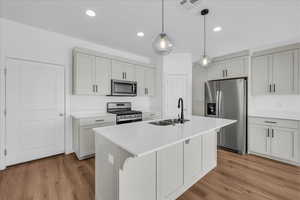 Kitchen featuring stainless steel appliances, decorative light fixtures, light wood-type flooring, a kitchen island with sink, and light stone counters