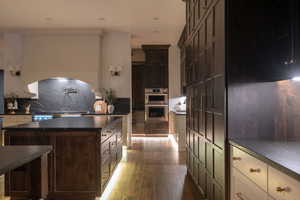 Kitchen with dark wood-type flooring, stainless steel double oven, dual tone cabinetry, tasteful backsplash, and dark stone counters