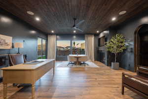 Home office featuring a lit fireplace, ceiling fan, arched walkways, light wood finished floors, and wooden ceiling