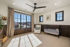 Bedroom featuring a nursery area, light colored carpet, a ceiling fan, a wainscoted wall, and wooden walls