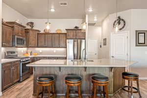 Kitchen featuring stainless steel appliances, a kitchen bar, a kitchen island with sink, decorative light fixtures, and light wood-style flooring