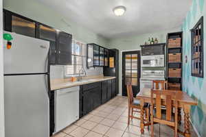Kitchen with glass fronted cabinets, white appliances, light countertops, dark cabinetry, and light tile patterned floors