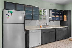 Kitchen featuring freestanding refrigerator, white dishwasher, glass insert cabinets, light countertops, and backsplash