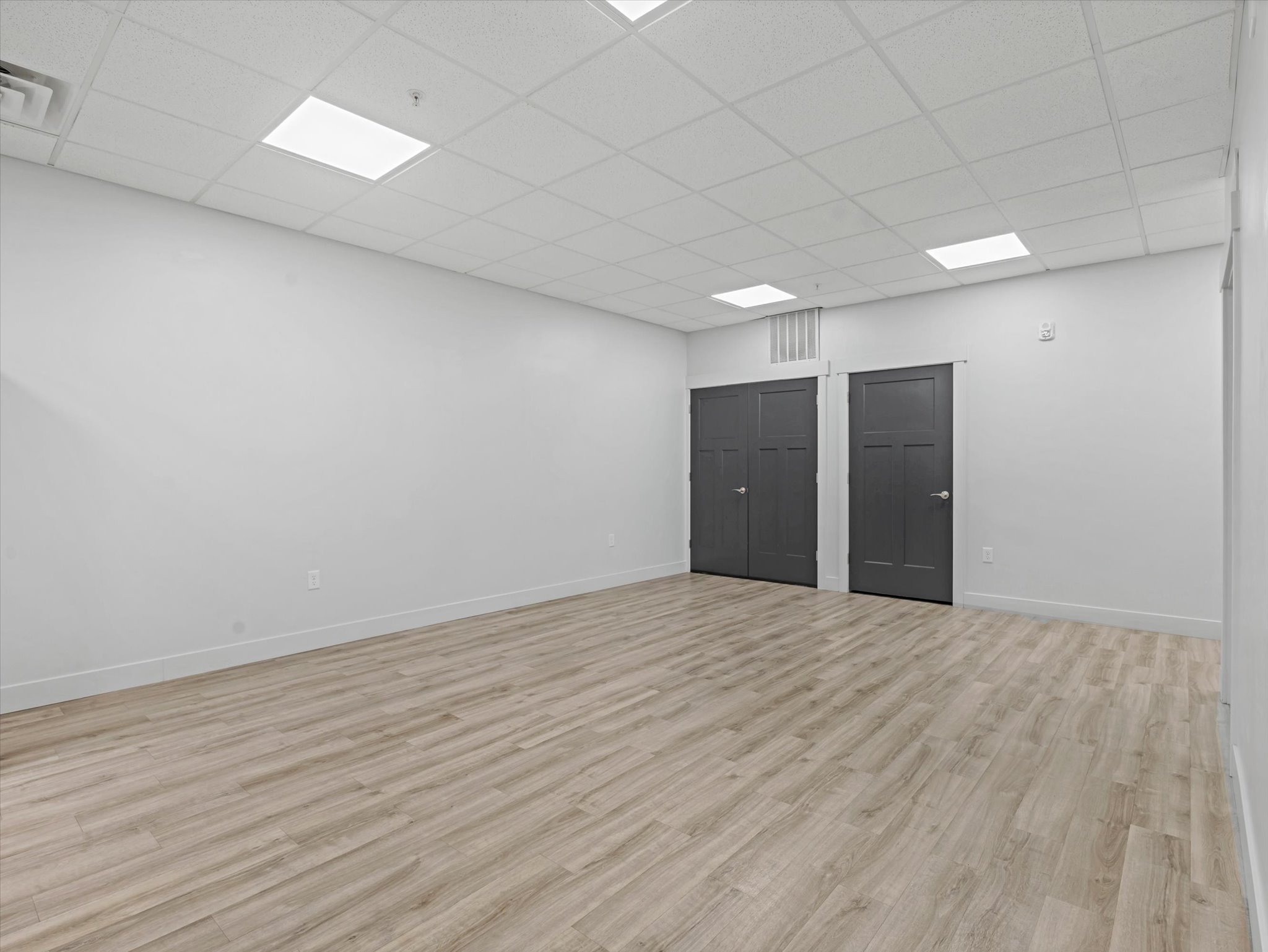 Spacious room featuring light wood-finish flooring, white walls, and a white grid-patterned drop ceiling with integrated rectangular lighting