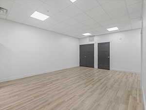 Spacious room featuring light wood-finish flooring, white walls, and a white grid-patterned drop ceiling with integrated rectangular lighting