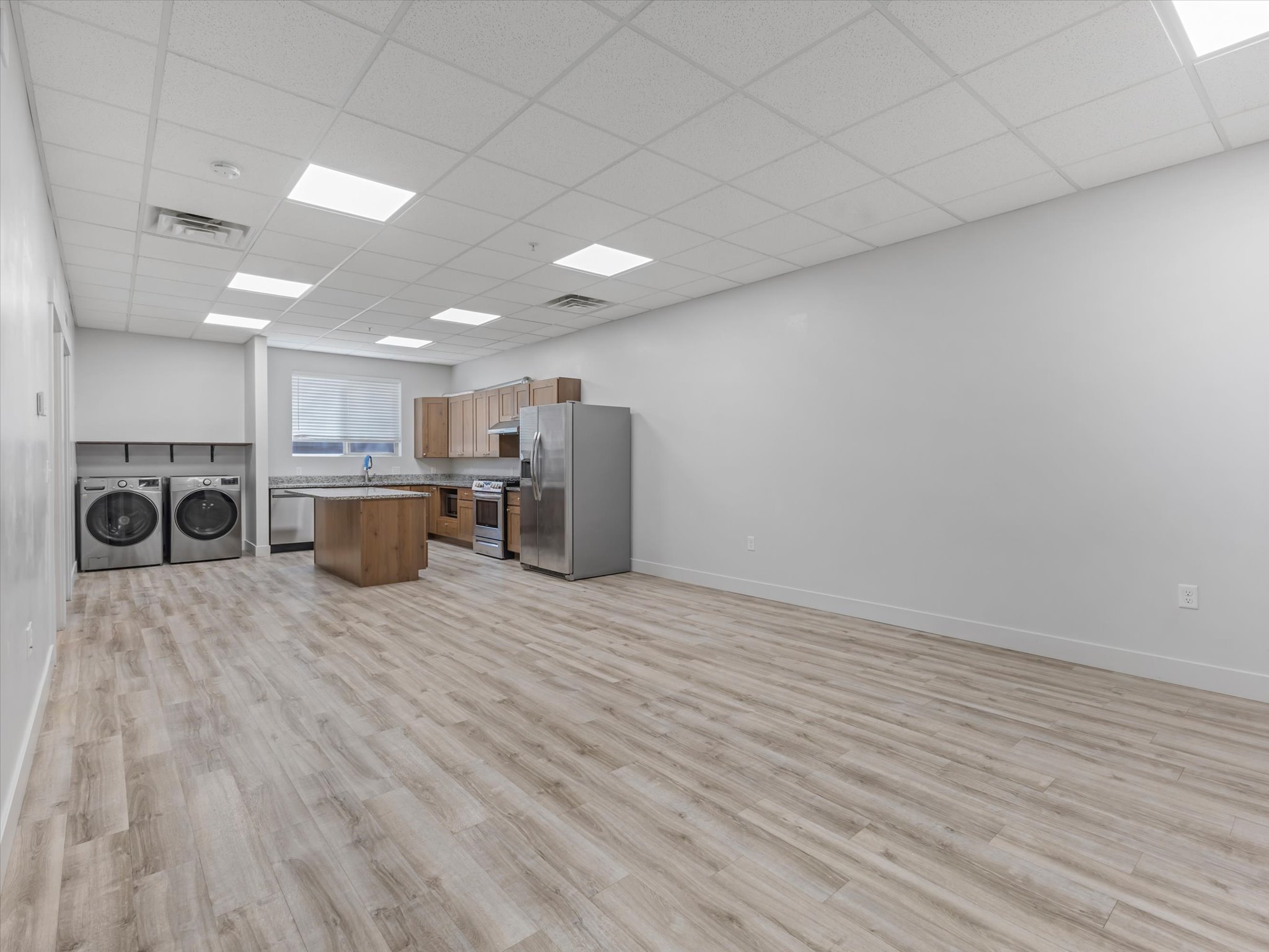 Open-concept living area featuring light wood-finish flooring, a kitchen with wood cabinetry and stainless steel appliances, a central kitchen island, and a dedicated laundry area with built-in shelving