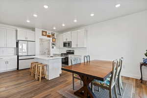 Kitchen featuring stainless steel appliances, dark wood-type flooring, white cabinetry, an island with sink, and a breakfast bar area