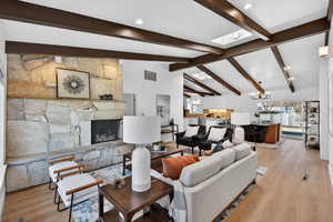 Gather in the Great Room, where soaring vaulted ceilings with exposed wood beams meet a grand stone fireplace. Natural light floods the space through dual skylights, highlighting the light wood-style flooring—the perfect centerpiece for both quiet eveni