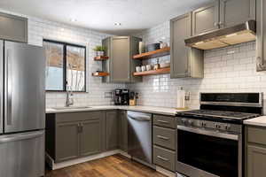 Kitchen featuring gray cabinetry, stainless steel appliances, open shelves, dark wood-style flooring, and backsplash