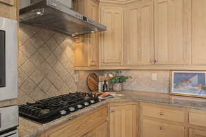 Kitchen with extractor fan, light stone counters, stainless steel appliances, backsplash, and light wood finish cabinetry