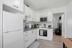 Kitchen featuring white appliances, light wood-type flooring, dark countertops, white cabinetry, and a textured ceiling