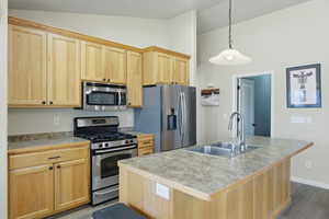 Kitchen featuring light wood finish cabinets, stainless steel appliances, a center island with sink, vaulted ceiling, and hanging light fixtures