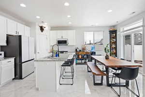 Kitchen featuring light stone countertops, freestanding refrigerator, a kitchen island with sink, white cabinets, and light marble finish flooring