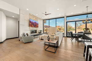 Living room with light wood-style flooring, a stone fireplace, floor to ceiling windows, a ceiling fan, and suspended lighting